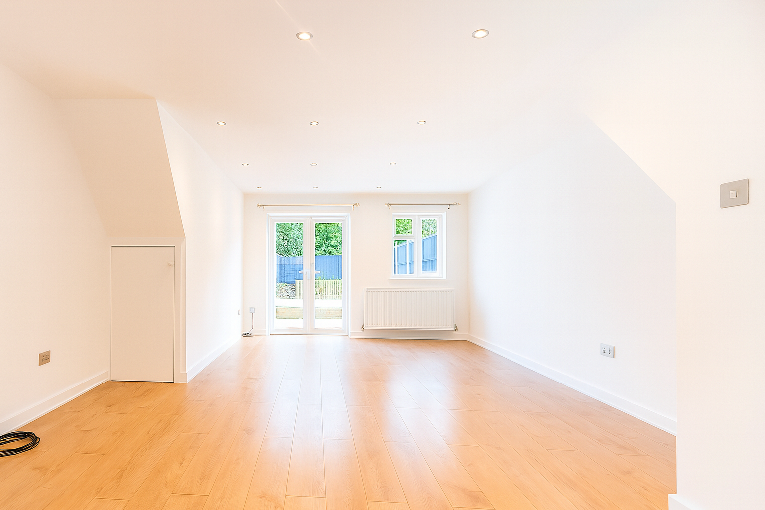 Bright renovated room with wooden flooring, recessed ceiling lights, double glass doors, and garden view through large windows.