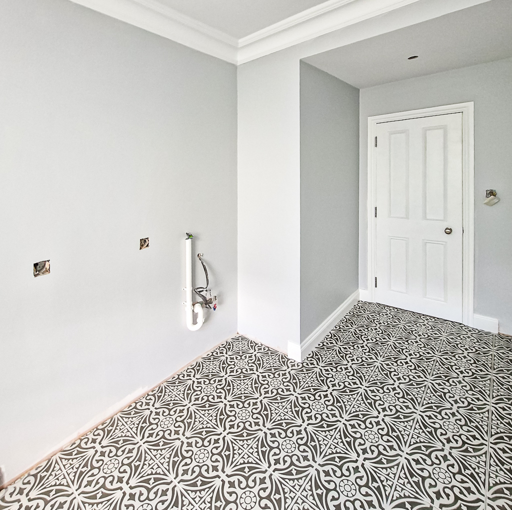 Utility room with decorative grey‑and‑white patterned floor tiles, light grey walls, white double doors, and exposed plumbing ready for installation.